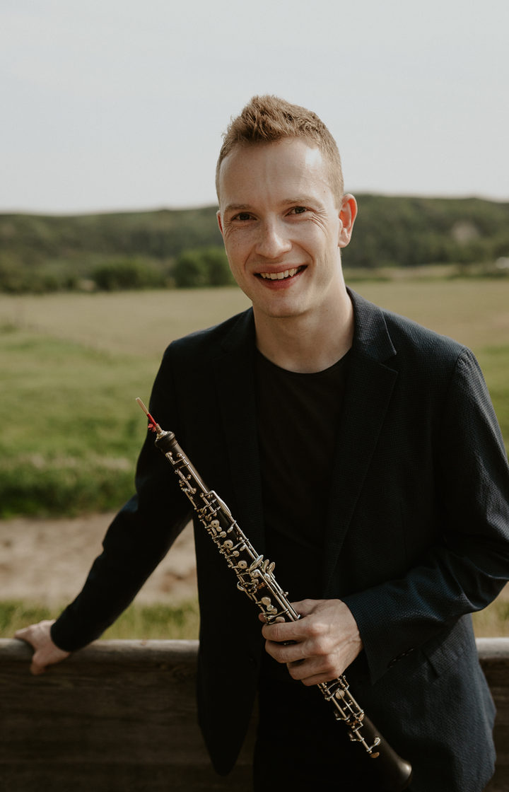 Aleh Remezau smiling, holding his oboe, leaning on a wooden fence in a field