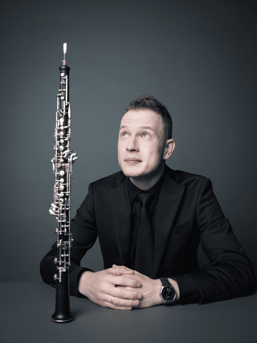 Aleh Remezau in a black suit at a table, looking up at his oboe standing beside him